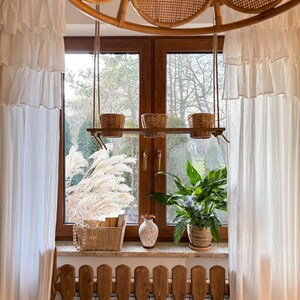 Könnte beinhalten: Ein Fenster mit weißen Vorhängen und einem Holzregal mit drei hängenden Körben. Das Regal ist über einem Holzheizkörper angebracht. Auf dem Fensterbrett steht eine Topfpflanze und auf dem Regal ein Weidenkorb mit Büchern.