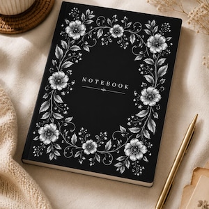 Op de afbeelding: Een zwart notitieboek met een witte bloemrijke randillustratie en het woord NOTEBOOK in het midden gedrukt. Het ontwerp van de kaft bevat gedetailleerde bladeren en bloesems in een ovaal kader met een decoratieve lijn onder de tekst.