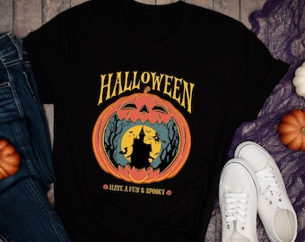 May include: Black Halloween t-shirt with a pumpkin graphic and the word "HALLOWEEN". The shirt is paired with blue jeans, white sneakers, and decorative pumpkins. The text "HAVE A FUN & SPOOKY" is also on the shirt.