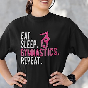 May include: Black t-shirt with the text "Eat. Sleep. Gymnastics. Repeat." in white with a pink silhouette of a gymnast performing a handstand.