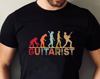 May include: A black t-shirt featuring a vintage-style graphic depicting the evolution of a guitarist. The graphic shows a series of figures progressing from a primitive ape to a modern-day guitarist holding a guitar. The text "GUITARIST" is printed below the graphic.