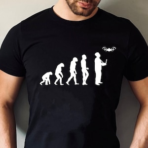 Może przedstawiać: Czarny T-shirt z białym nadrukiem przedstawiającym ewolucję człowieka, kończącą się na człowieku trzymającym drona. Na koszulce widnieje napis "Drone Evolution".