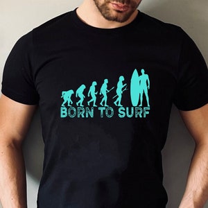 May include: A black t-shirt with a graphic of the evolution of humans ending with a surfer holding a surfboard. The text "BORN TO SURF" is printed below the graphic.