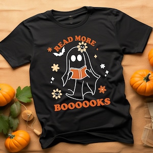 Op de afbeelding: Zwart t-shirt met een spookafbeelding die een boek leest, omringd door bloemen en sterren. De oranje tekst luidt "READ MORE BOOOOKS". Pompoenen zijn rond het shirt geplaatst.