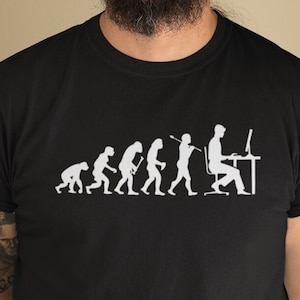 Peut inclure: Un t-shirt noir avec un graphique blanc représentant l'évolution des humains se terminant par une personne assise devant un ordinateur. La personne porte un casque et regarde l'écran d'un ordinateur.