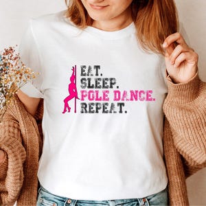 Könnte beinhalten: Weißes T-Shirt mit dem Text "Eat. Sleep. Pole Dance. Repeat." in schwarzer und rosa Schrift. Links vom Text befindet sich eine rosa Silhouette einer Frau, die Pole Dance tanzt.