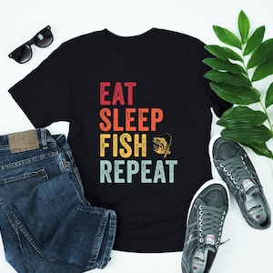 Puede incluir: Camiseta negra con un gráfico de estilo vintage que dice "Eat Sleep Fish Repeat" con una silueta de un pez y una caña de pescar.