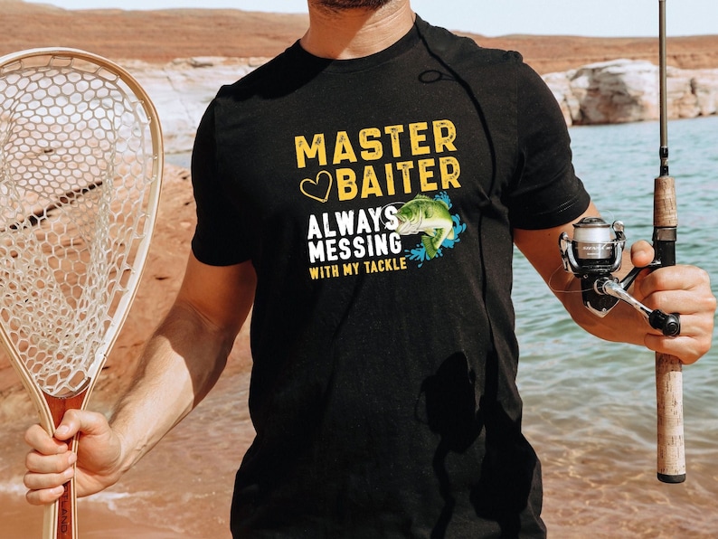 May include: Black t-shirt with the text "MASTER BAITER" in yellow and white, a heart symbol, and a graphic of a fish. The shirt also says "ALWAYS MESSING WITH MY TACKLE". A fishing rod and net are also visible.