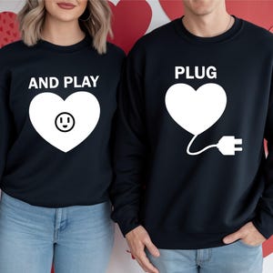 May include: Two navy blue sweatshirts with white heart graphics. One says "AND PLAY" with a smiley face in the heart, the other says "PLUG" with a plug graphic. The sweatshirts are worn with blue jeans.