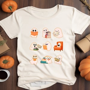 May include: Cream-colored t-shirt featuring a collection of cartoon ghosts reading books. The ghosts are in various poses, some with glasses, and one is seated in an orange armchair. The design has a Halloween or fall theme.