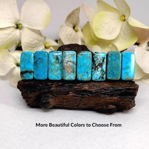 May include: Eight rectangular turquoise stones with varying blue hues and dark brown matrix patterns arranged in a row on a dark textured wooden base. The text "More Beautiful Colors to Choose From" is printed below the wooden base.