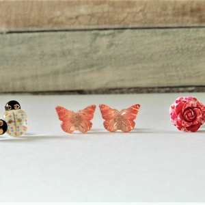 May include: Three pairs of earrings featuring different designs: a pair of ladybug earrings with pink and blue polka dots, a pair of butterfly earrings with a red and gold design, and a pair of rose earrings with a white and red design.