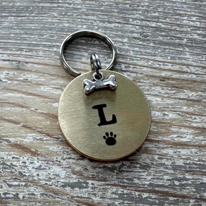 May include: Gold dog tag with a bone charm and the letter L engraved on it. The tag has a paw print engraved below the letter.