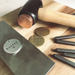 May include: A close-up of a metal stamping kit with three round metal tags. The tags are engraved with the words "benjie", "dachshund mama", and "billiest mom". The kit also includes a black rubber mallet and several metal stamping tools.
