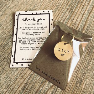 May include: A gold-colored metal dog tag with the name "LILY" engraved on it. The tag is attached to a silver key ring and is packaged in a clear plastic bag with a brown paper backing that says "ink and doodle".