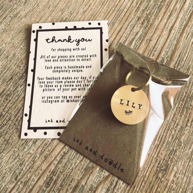 May include: A gold-colored metal dog tag with the name "LILY" engraved on it. The tag is attached to a silver key ring and is packaged in a clear plastic bag with a brown paper backing that says "ink and doodle".