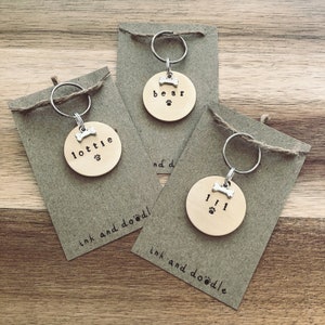 May include: Three personalized dog tags with bone charms and paw prints. The tags are made of wood and have the names "Lottie", "Bear", and "Lil" engraved on them. The tags are attached to key rings and are displayed on brown paper cards with the text "ink and doodle" printed on them.