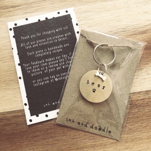 May include: A gold-colored metal dog tag with the word "bear" engraved on it. The tag has a small bone charm attached to it. The tag is hanging from a silver key ring. The tag is on a brown background with the text "ink and doodle" printed on it.