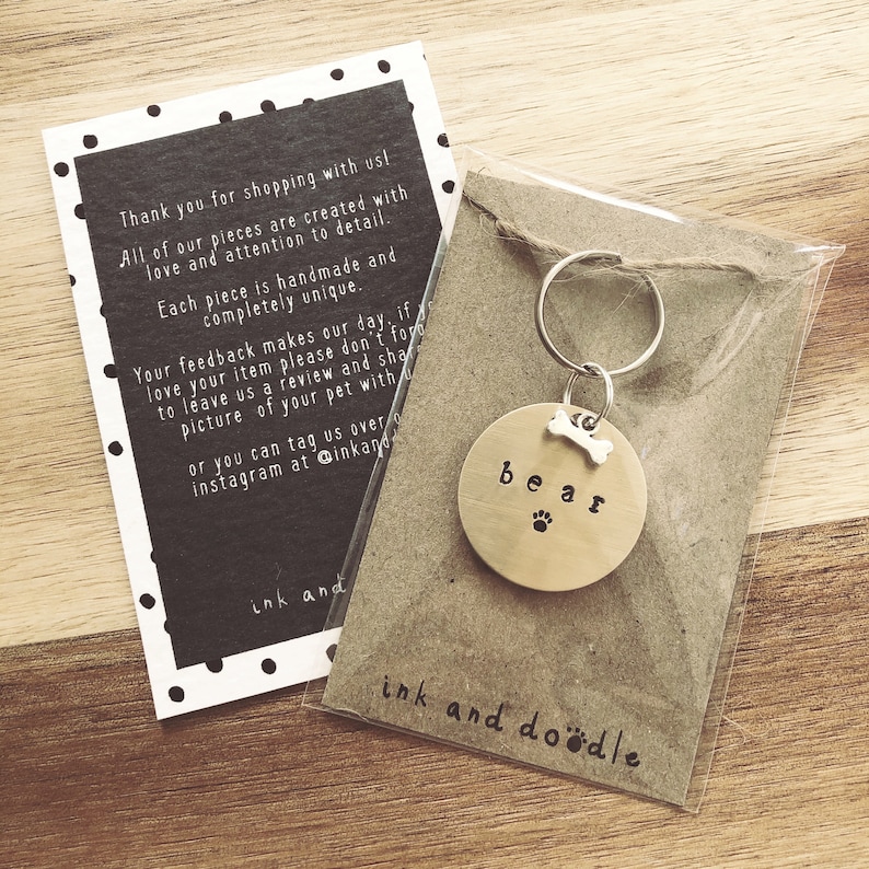 May include: A gold-colored metal dog tag with the word "bear" engraved on it. The tag has a small bone charm attached to it. The tag is hanging from a silver key ring. The tag is on a brown background with the text "ink and doodle" printed on it.
