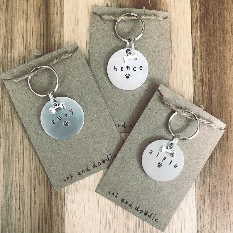 May include: Three personalized dog tag keychains with the names "Ruby", "Bruce", and "Alfie" on them. Each tag has a small bone charm and a paw print. The tags are displayed on brown card stock with the text "ink and doodle" printed on them.