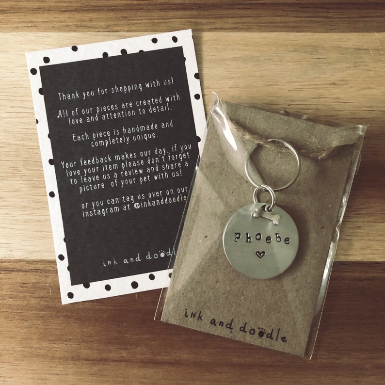 May include: A silver dog tag with the name "Phoebe" engraved on it, with a small heart and bone symbol. The tag is attached to a silver key ring and is packaged in a brown paper bag with the words "ink and doodle" printed on it.