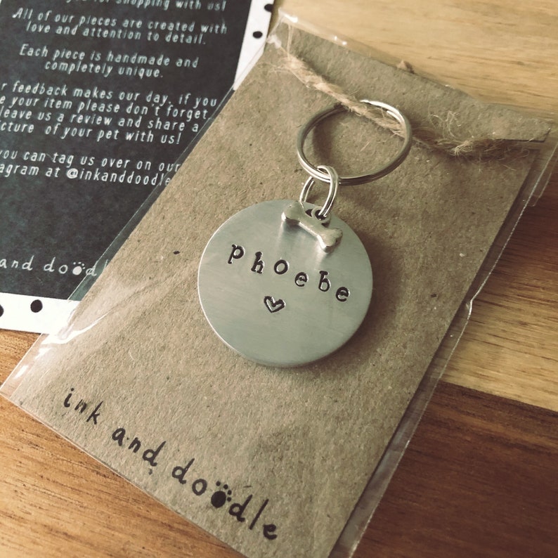 May include: A silver dog tag with the name "Phoebe" engraved on it, a small heart, and a bone charm. The tag is attached to a silver key ring and is displayed on a brown paper card with the text "ink and doodle" written on it.