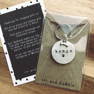 May include: Silver dog tag with the name "bruce" engraved on it, a bone and paw print design, and a silver key ring. The tag is in a clear plastic bag with a brown paper backing that says "ink and doodle".