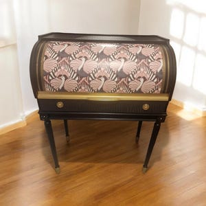May include: A black and gold vintage cabinet with a patterned front featuring pink and white swan designs. The cabinet has a curved top, two small drawers, and slender legs with gold accents. It is set on a wooden floor.