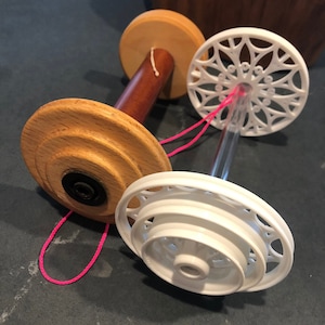 Puede incluir: Dos carretes de hilo de madera con una cuerda rosa atada. Un carrete es de madera natural con una rueda de plástico blanco. El otro carrete es de madera marrón con una rueda de plástico blanco con un diseño decorativo.