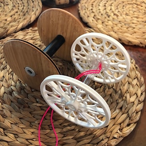 Puede incluir: Un enhebrador de hilo blanco con dos carretes, uno de madera y otro de plástico. Hilo rosa enhebrado a través del enhebrador.