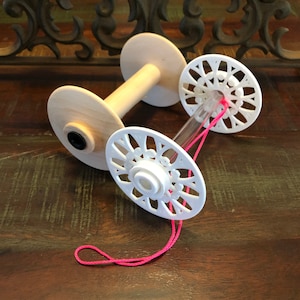 Puede incluir: Un torno de madera para hilados con dos bobinas de plástico blanco. El torno tiene un tubo central de plástico transparente y un hilo rosa está envuelto alrededor de las bobinas y el tubo.