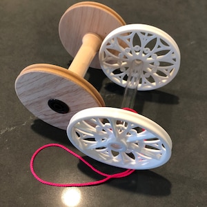 Puede incluir: Un torno de madera para hilados con dos ruedas blancas decorativas de trabajo abierto. Una cuerda rosa está unida al torno.