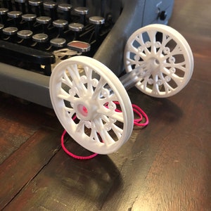 Puede incluir: Dos carretes de plástico blanco con un diseño decorativo, conectados por una cuerda rosa. Los carretes están sobre una superficie de madera.