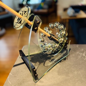 Pode incluir: Um enrolador de bobina de acrílico transparente com um pino de madeira e detalhes decorativos. O enrolador tem uma base com o texto "WWW.BOBBINEXCHANGE.COM". O design inclui um elemento circular com pequenas esferas prateadas.