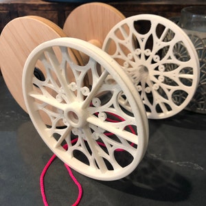 Puede incluir: Un conjunto de ruedas decorativas de madera y blancas. Las ruedas blancas tienen un diseño abierto y estampado y están conectadas con una cuerda rosa brillante. Las ruedas de madera son macizas y redondas.