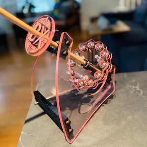 Puede incluir: Un bobinador de acrílico rosa con una clavija de madera y detalles decorativos. El bobinador tiene una base transparente y un diseño circular con el texto "DIY" en el lateral. La dirección del sitio web www.bobbinexchange.com es visible en la base.