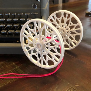 Pode incluir: Dois carretéis de plástico branco com um design decorativo e intrincado. Os carretéis estão conectados por um fio rosa.