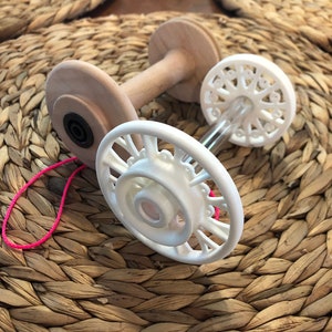 Puede incluir: Un enhebrador de lana de madera blanca con dos ruedas de plástico blanco. El enhebrador está sobre una superficie de mimbre marrón tejido.