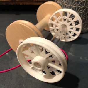 Puede incluir: Un enhebrador de lana de madera blanca con dos carretes y una cuerda roja. El enhebrador tiene un diseño decorativo en las ruedas.