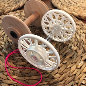 Puede incluir: Un enhebrador de lana de madera blanca con dos carretes blancos y un hilo rosa atado a uno de los carretes. El enhebrador está sobre una superficie de mimbre marrón tejido.