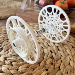 Puede incluir: Dos bobinas blancas impresas en 3D con un diseño calado decorativo. Las bobinas están conectadas por un tubo transparente. Una bobina tiene el texto "BOBBIN EXCHANGE" impreso. Las bobinas están sobre una superficie tejida y texturizada.