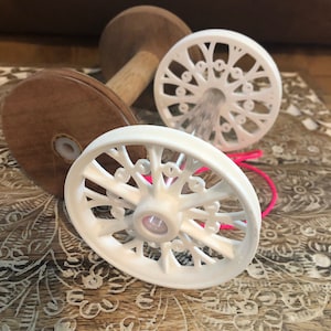 Puede incluir: Dos carretes de madera blancos con diseños intrincados, un carrete está encima del otro, con una cuerda rosa envuelta alrededor de los carretes.