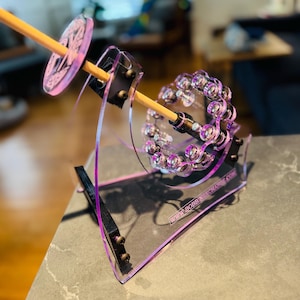 Pode incluir: Uma roda de fiar em acrílico transparente com detalhes roxos. A roda tem um fuso de madeira e um elemento circular decorativo. A base tem o texto "WWW.BOBBINNEWCHANGE.COM".