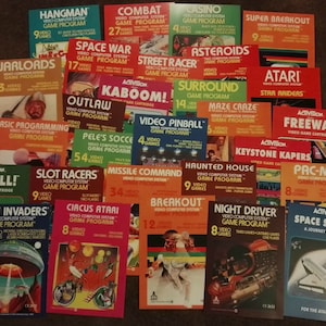 Puede incluir: Una colección de cartuchos de juegos de video Atari vintage, incluyendo clásicos como Pac-Man, Space Invaders y Pitfall!. Los cartuchos están dispuestos en una cuadrícula sobre una superficie marrón. Cada cartucho presenta una caja colorida con un título de juego, una descripción y una ilustración.