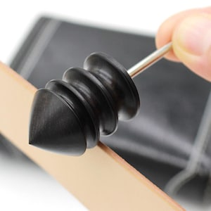 Puede incluir: Una herramienta de artesanía en cuero negra con una punta cónica y una serie de crestas redondeadas. La herramienta está sujeta por una mano y se coloca sobre un trozo de cuero marrón claro. La herramienta tiene un eje de metal plateado.