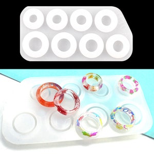 May include: A white silicone mold with eight ring-shaped cavities. The mold is filled with colorful resin rings with floral and geometric designs.