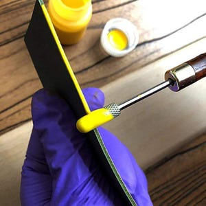 以下が含まれることがあります： 革細工プロジェクトのクローズアップ。黒い革片に、特殊な工具を使って黄色のエッジペイントを塗布しています。黄色の塗料のボトルと綿棒が背景にあります。