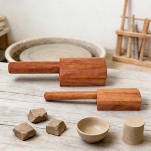 Op de afbeelding: Twee houten hamers met een gladde, roodbruine nerf, vergezeld van drie kleine, onregelmatige stukjes klei, een kleine beige keramische kom en een massieve beige cilindrische kleivorm, gerangschikt op een lichtgekleurd houten oppervlak.