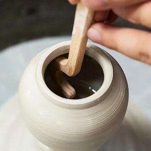 May include: A light-colored ceramic vessel on a pottery wheel being shaped from the inside with a wooden L-shaped rib tool. The clay pot has a smooth, rounded body and a circular opening at the top, with visible horizontal lines from the wheel.