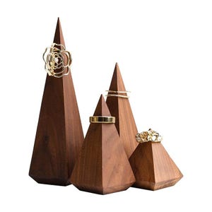 May include: Four dark brown wooden ring holders in varying heights and pyramid shapes. Each wooden cone displays a gold-toned ring, including a floral design, a plain band, a thin stacked band, and a textured ring, arranged in a cluster.